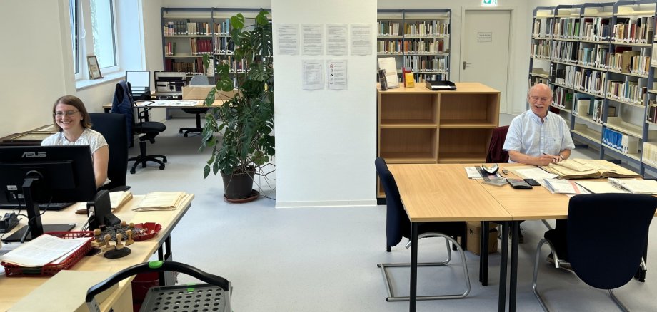La photo montre une vue de la nouvelle salle de lecture avec des étagères et des tables. À gauche de la photo, le Dr Svenja Müller est assise à un bureau. À droite, Dieter Jerusalem est assis à une table avec des documents anciens.