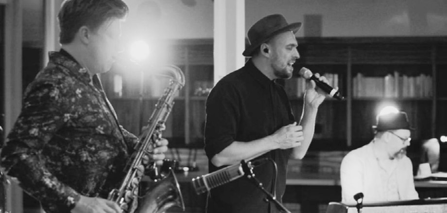 La photo en noir et blanc montre au premier plan un saxophoniste, derrière lui Max Mutzke avec un micro en train de chanter et Matti Klein en arrière-plan au clavier.
