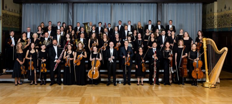 La photo montre les membres de l'orchestre d'État avec leurs instruments, disposés en quatre rangées ascendantes dans une salle de réception ornée de lustres.