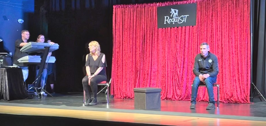 La photo montre une actrice et un acteur du théâtre Requisit, assis sur des chaises devant un rideau rouge sur la scène du théâtre municipal. A gauche de l'image, on voit encore deux techniciens avec leur équipement.