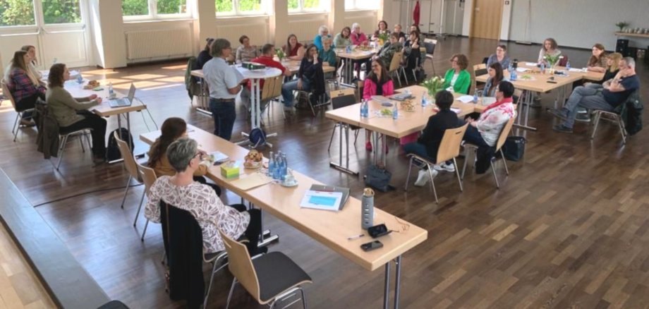 La photo montre les participants à la conférence de direction, assis à des tables dans l'aula de Göttenbach. Au milieu d'eux se trouve Heinz Müller qui fait un exposé.