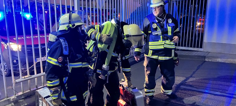 La photo montre deux pompiers équipés de masques respiratoires et d'autres équipements de secours. Un chef d'équipe leur donne des instructions.