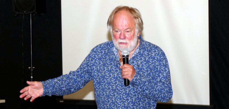 La photo montre l'auteur se tenant devant un écran avec un microphone pendant sa conférence.