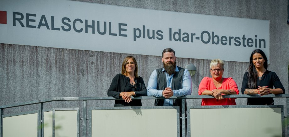 La photo montre les personnes susmentionnées, alignées et appuyées sur une balustrade devant un panneau portant l'inscription "Realschule plus Idar-Oberstein".