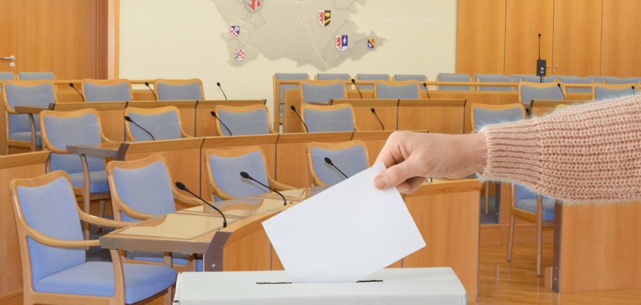 Das Foto zeigt einen Arm, der einen Stimmzettel in eine Wahlurne steckt. Im Hintergrund ist der Sitzungssaal der Stadtverwaltung Idar-Oberstein zu sehen.