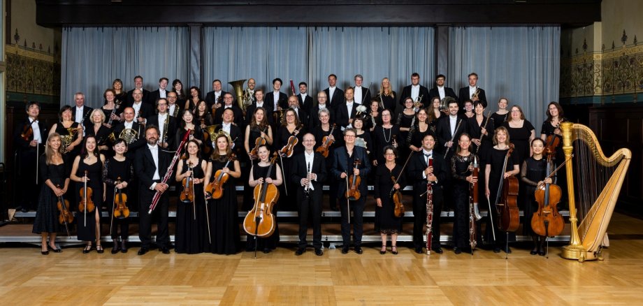 Das Foto zeigt die Orchestermitglieder in vier aufsteigenden Reihen in einem Festsaal.