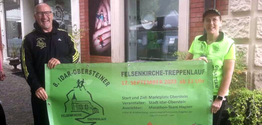 Das Foto zeigt das Ehepaar Rainer und Ilonka Hagner. Sie halten ein Banner mit Werbung für den 8. Idar-Obersteiner Felsenkirche-Treppenlauf.