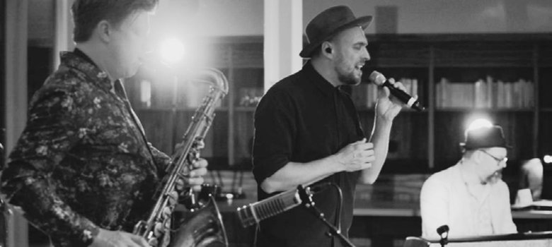 Das Schwarzweiß-Foto zeigt im Vordergrund einen Saxophonisten, dahinter Max Mutzke mit dem Mikro beim Singen und Matti Klein im Hintergrund am Keyboard.