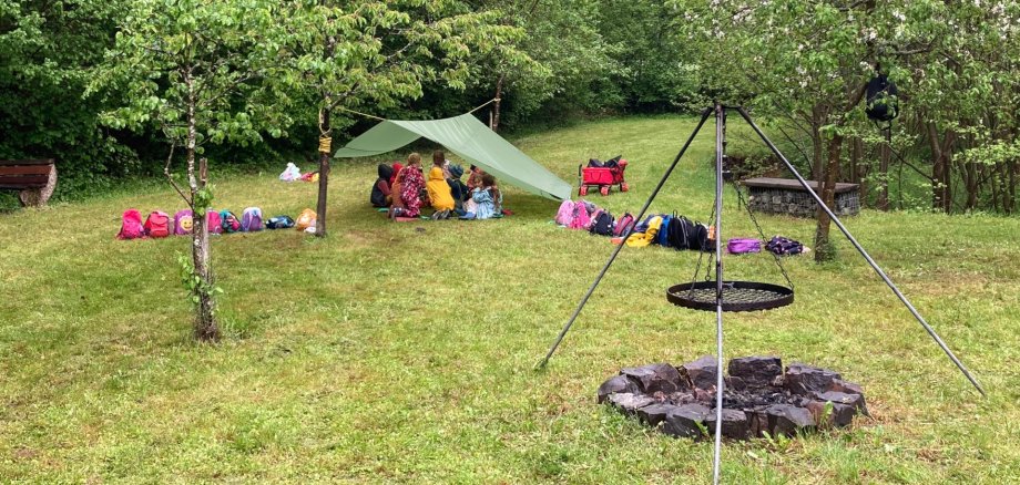 Das Foto zeigt im Hintergrund die Kinder, die unter einer Zeltplane auf einer Wiese sitzen. Im Vordergrund ist ein Dreibein mit Schwenkrost zu sehen.