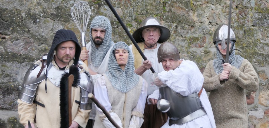 Das Foto zeigt die Schauspieler in Ritterkostümen mit Waffen vor einer Steinwand.