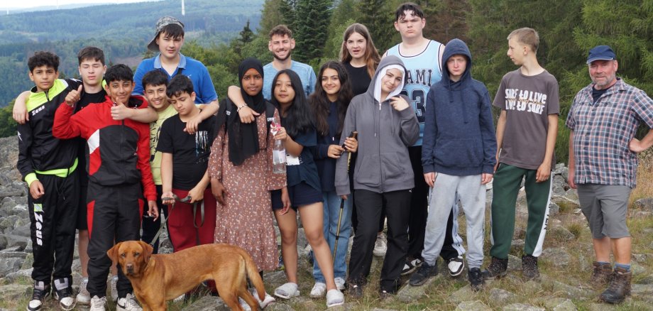 Das Foto zeigt die Schüler und ihre Begleitpersonen. Sie stehen in zwei Reihen nebeneinander vor einem Waldrücken und blicken in die Kamera. Vor der Gruppe steht noch ein Hund.