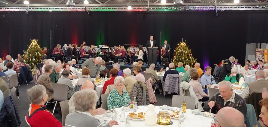 Das Foto zeigt einen Blick in die Messehalle mit zahlreichen Gästen an Tischen. Auf der Bühne im Hintergrund sitzen die Mitglieder des Musikvereins Tiefenstein. Am Mikrofon steht Oberbürgermeister Frühauf, neben ihm Karl-Heinz Totz.