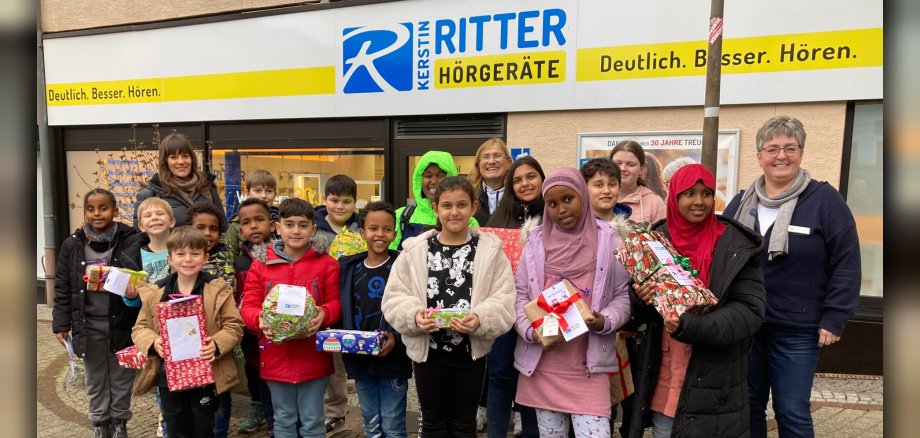 Das Foto zeigt die Kinder und Betreuer der Spiel- und Lernstube sowie die Mitarbeiterinnen der Filiale. Sie stehen vor dem Geschäft und die Kinder haben die Geschenke in den Händen.