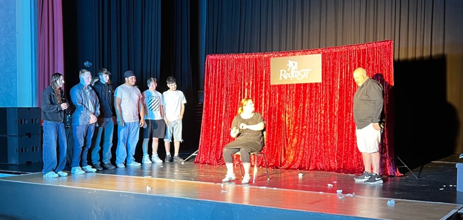 Das Foto zeigt einen Blick auf die 'Bühne des Stadttheaters. Vor einem roten Vorhang sieht man zwei Darsteller des Theaters Requisit. Daneben stehen sechs Schüler und blicken auf die Darsteller.