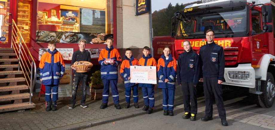 Das Foto zeigt eine Abordnung der Jugendfeuerwehr, die gemeinsam mit Bäckermeister Michael Merz vor dem Bäckerladen steht. Sie stehen nebeneinander, Bäcker Merz hält einen Korb mit Brezeln in der Hand, zwei Mitglieder der Feuerwehr einen großen Spendenscheck. Dahinter ist ein Feuerwehrauto zu sehen. 