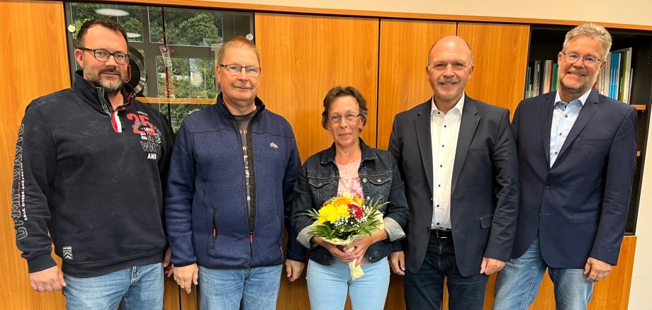 Das Foto zeigt fünf Personen, die nebeneinander vor einer Schrankwand stehen und in die Kamera schauen. Frau Di Cato steht mit einem Blumenstrauß in den Händen in der Mitte.