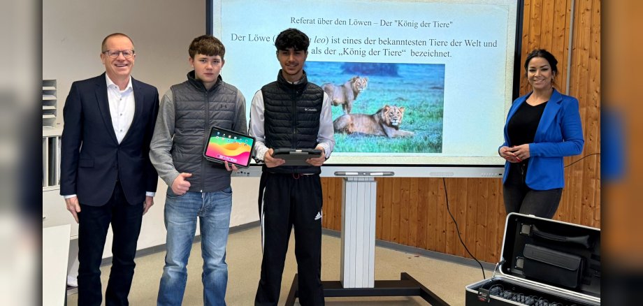 Das Foto zeigt die beiden genannten Personen, die gemeinsam mit zwei Schülern vor einem Whiteboard stehen. Die Schüler haben noch jeweils ein Tablet in der Hand.